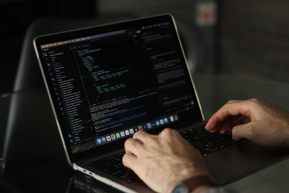 A close-up view of a person's hands typing on a laptop keyboard, with the screen displaying lines of programming code on a dark background. The laptop is placed on a smooth, reflective surface, possib