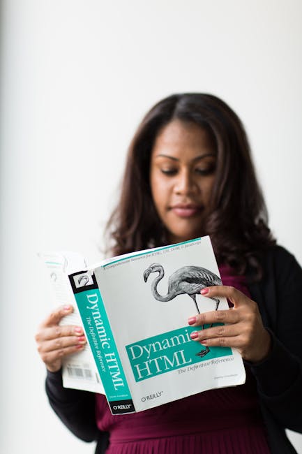 A woman with medium-length dark brown hair, wearing a black blazer over a maroon top, is holding a book titled 'Dynamic HTML' by O'Reilly. She is standing indoors against a plain white background, foc